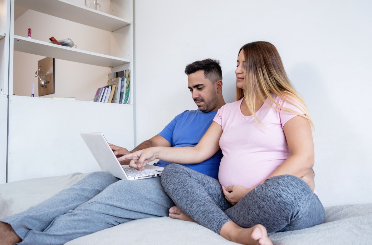 Couple watching laptop together, partner pointing at screen as pregnant woman listens