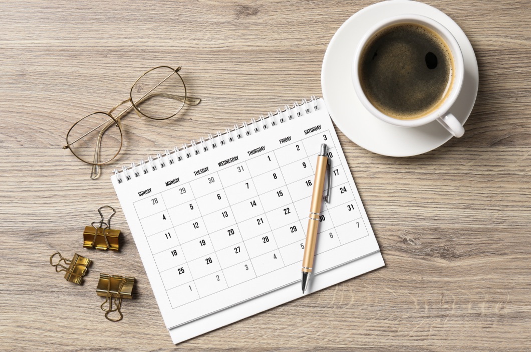 Calendar flat-lay with reading glasses, pen, and a cup of coffee on warm wooden surface