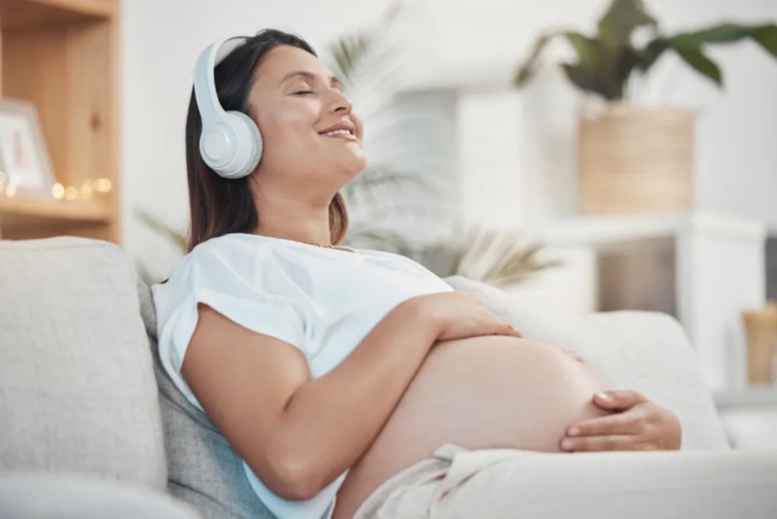 Pregnant woman reclined on a sofa, listening to audio through headphones with eyes closed