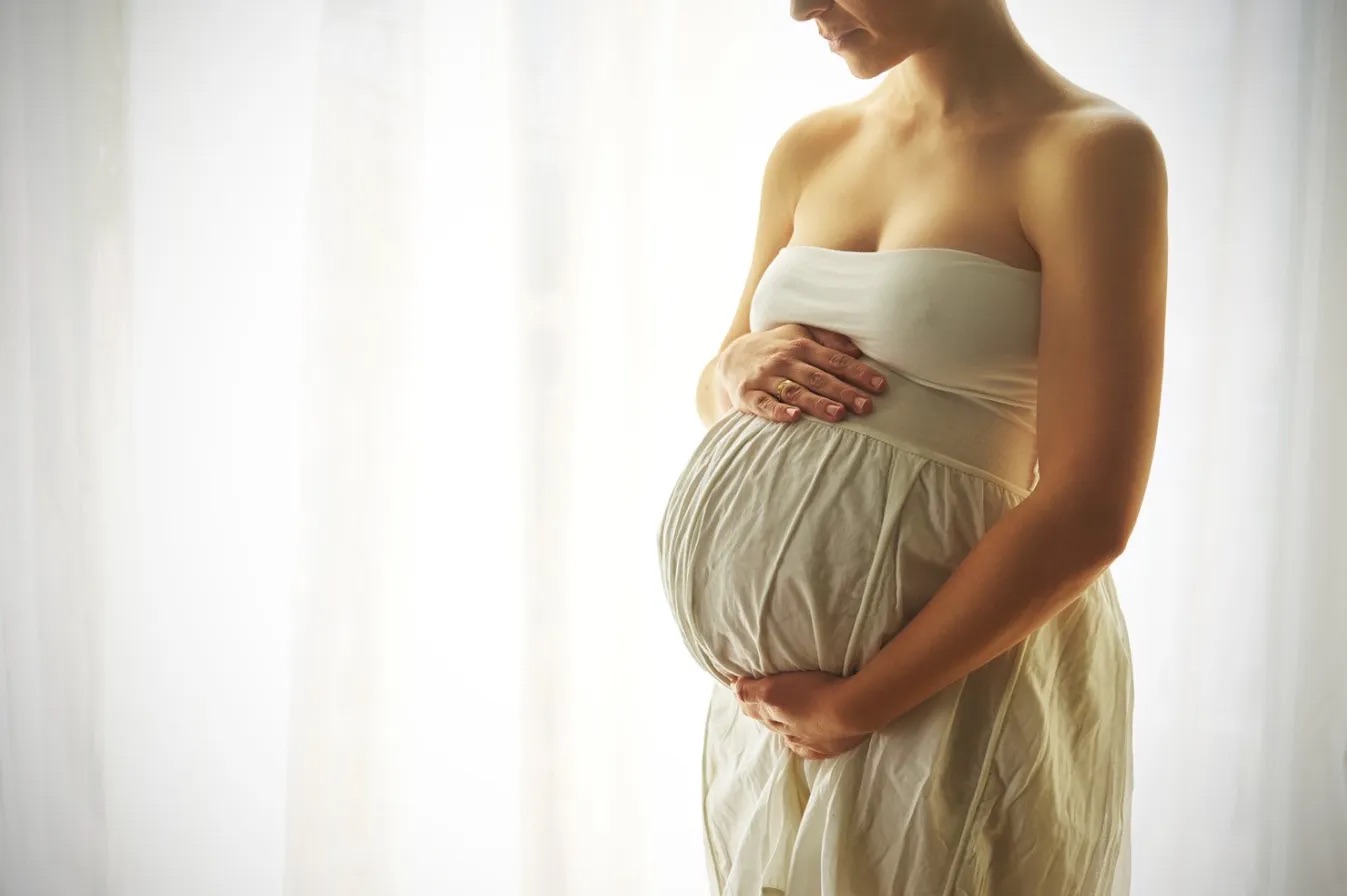 Pregnant woman in a soft cream dress standing by a window in calm natural light