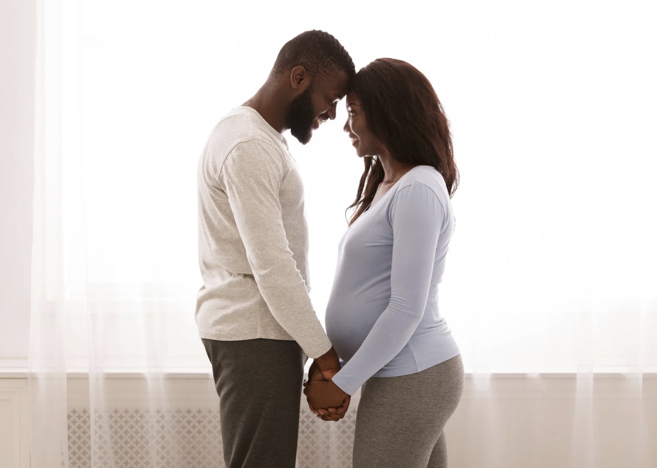 Pregnant woman and her partner standing forehead-to-forehead, holding hands by a window