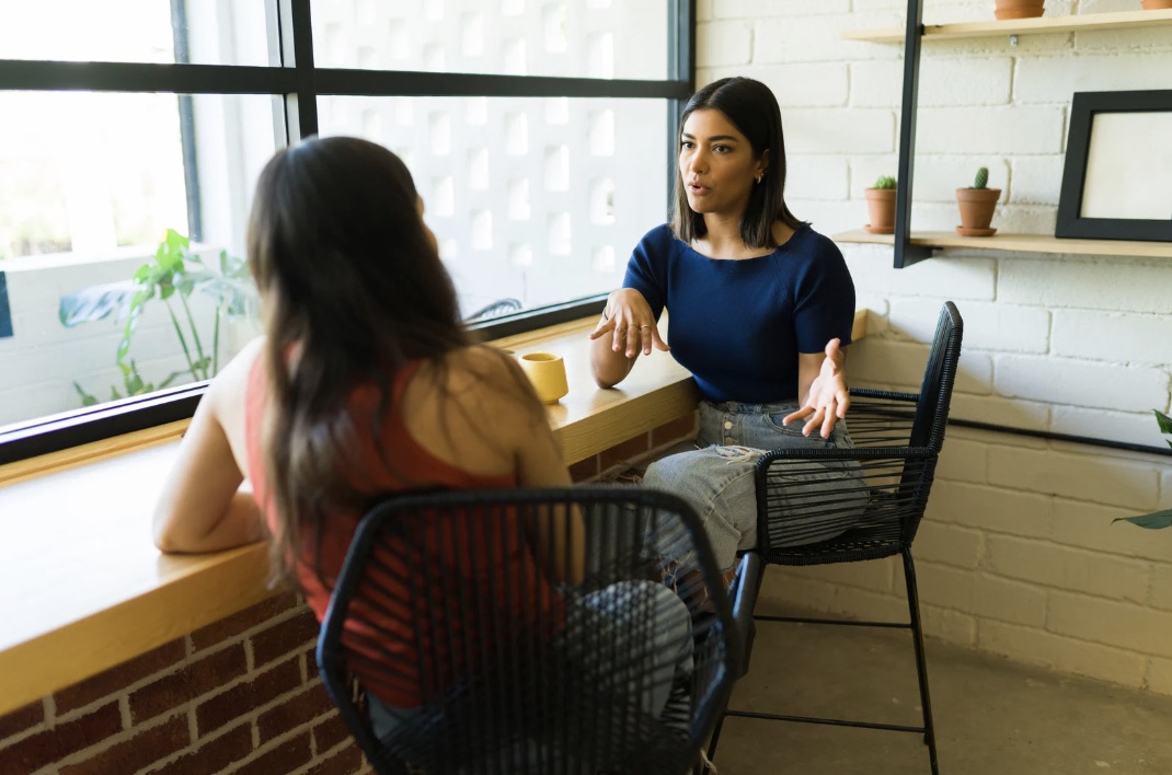 Two women in deep conversation across a wooden café counter, engaged dialogue