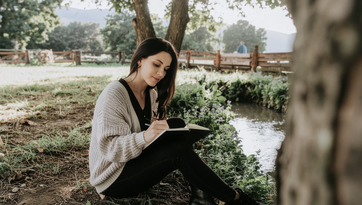 Woman in cream cardigan writing in journal outdoors by a stream, warm golden light