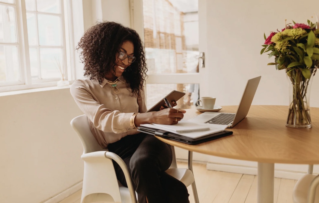 Smiling woman at a wooden table with laptop, tablet and flowers, engaged in her work