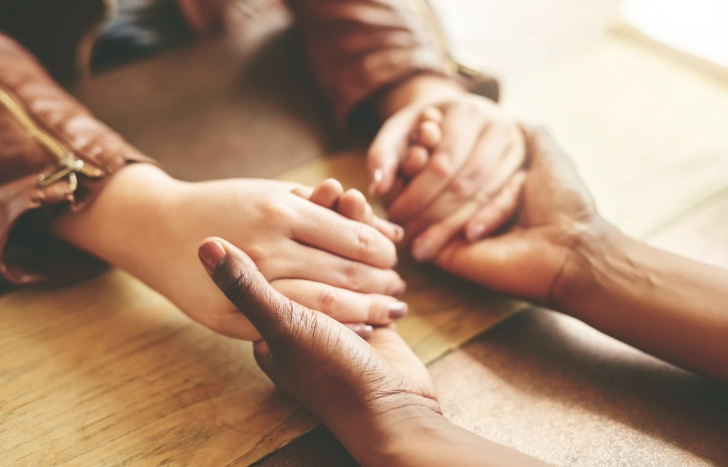 Two pairs of hands clasped together on a wooden table in warm golden light, gentle support